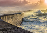 Waves crash against the concrete wall of the harbor at sunset.