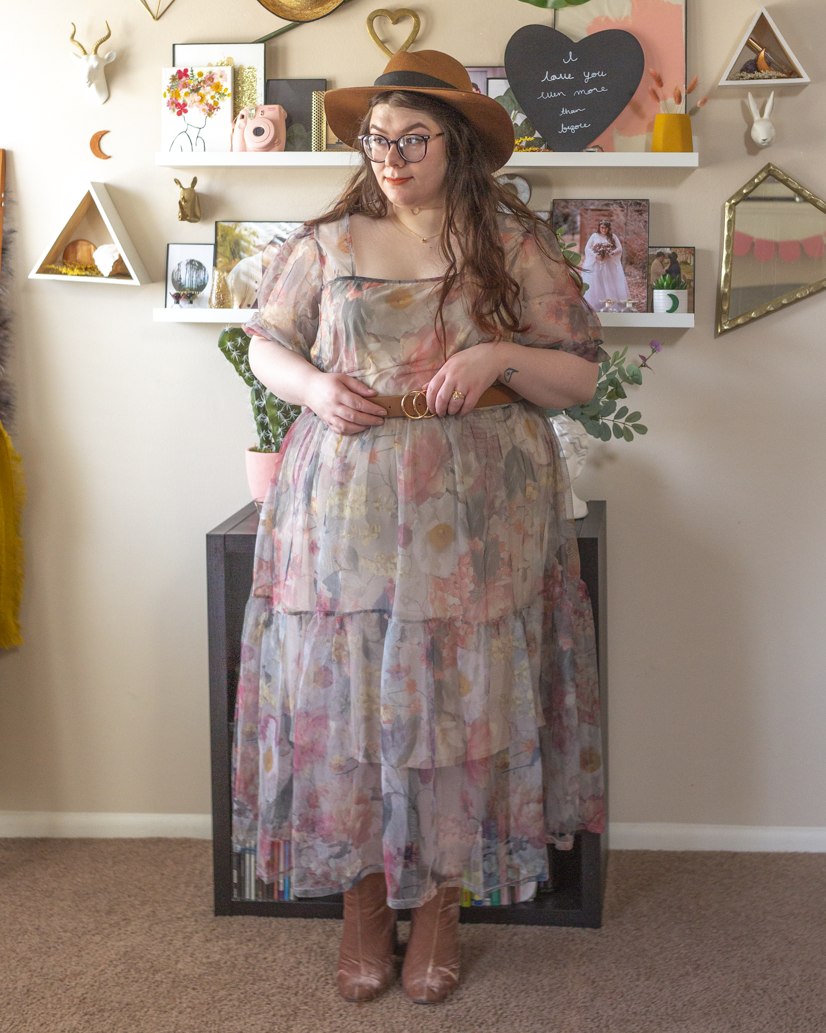 An outfit consisting of a brown wide brim fedora floppy hat, a beige square neck puffy sleeve with muted floral print, belted with a brown belt and pastel pink suede boots.