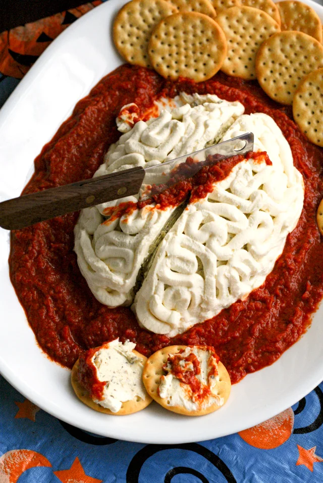 A cheese ball on the shape of a brain with a knife stuck in the top and tomato sauce around the knife and around the cheese ball.