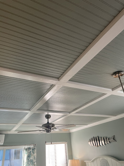 Picture of the ceiling which is made of wood, painted a blueish gray with white beams making a large square shaped design. There is a ceiling fan above the bed.