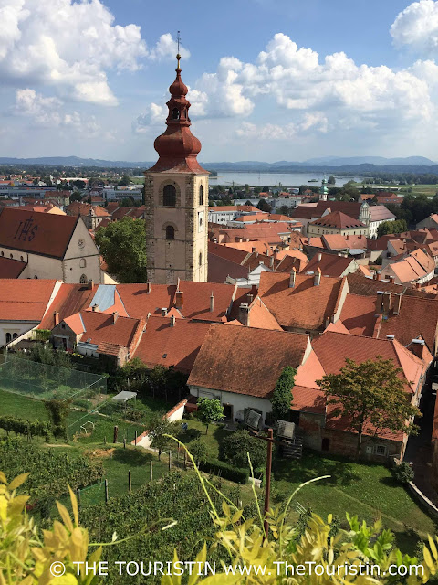 A tall red-roofed church tower in the midst of a village with small red-roofed period houses by a lake, surrounded by lush green hills, under a blue sky.