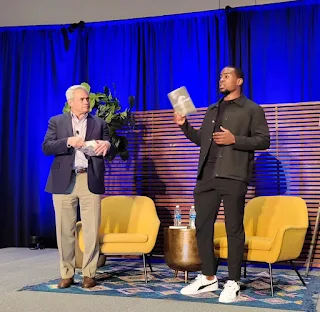 two men standing on stage with one holding a book