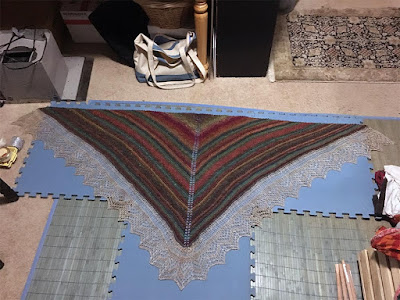 A large pink, teal, green, and gold shawl with chevron stripes and a wide white lace border, pinned out on a tee shape of blue interlocking foam floor pads, in a very cluttered room. There's stuff encroaching from all edges of this photo.