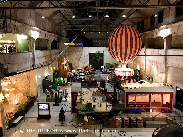 A white and red life-sized hot air balloon in a factory with concrete walls decorated with huts, plants, and lights.