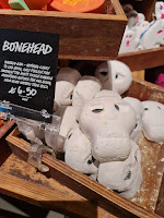 A photo of a light brown square box containing a bunch of white skull shaped bubble bars with holes to make a skulls face next to a black rectangular card that says bonehead bubble bar in white font on a light brown rectangular table on a bright background