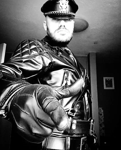 Black and white photograph of leather daddy pointing at camera wearing full gear including padded shoulder biker jacket