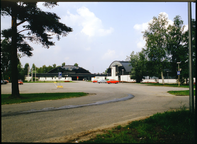 Uimahalli- ja urheilutalorakennukset kuvattuna parkkipaikan suunnasta sulan aikaan. Rakennusten edessä parkkeerattuina erivärisiä ja -merkkisiä autoja.