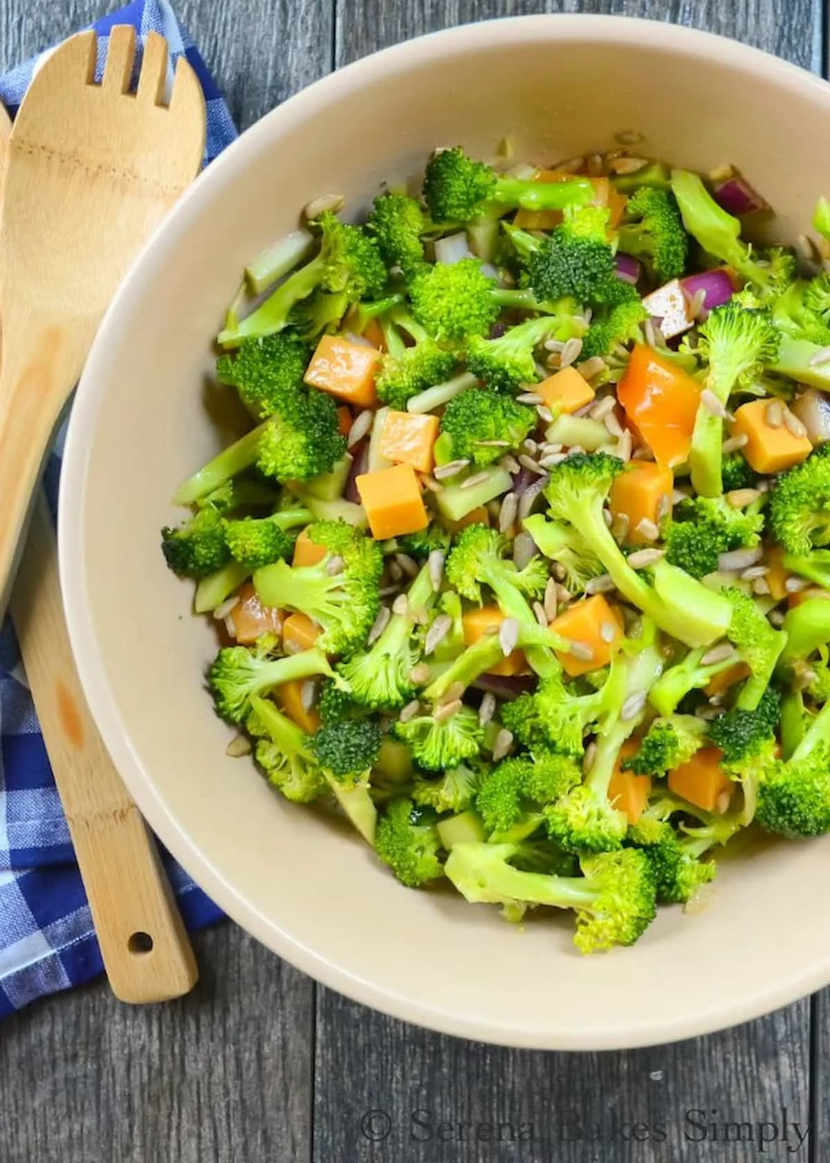 A down shot of Broccoli Salad in a cream colored bowl with wooden serving utensils to the left side of photo.