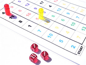 na zdjęciu jest plansza z kolorowymi liczbami podzielonymi na sekcje, które wyznaczają tor wyścigu, na planszy stoją pionek czerwony i żółty a obok leża trzy kostki z wynikami 6,3,3