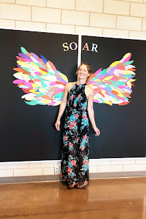 Alexia standing beside a poster wall in the high school, black background with multi colored paper wings and the word SOAR above