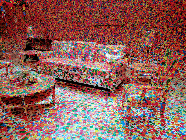 Photo of Yayoi Kusama's obliteration room. A room comprised entirely of white walls, white furniture and white objects. Guests to the show cover the objects with polka dot stickers on the was out. This flattens the space and create an illusion of disappearing objects.