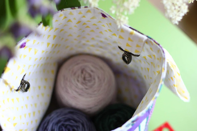 Interior view of a project bag lined with yellow triangle fabric. Two loops hanging into the bag from the top edge have bronze swivel snaphooks to be used as yarn guides. Three cakes of yarn sit at the bottom of the bag.