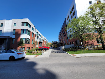 Looking between two new buildings along a block of street where the adjacent sidewalk is broken up by a driveway and TWSI on either end of the sidewalk. (Rochester Street, east side, between Balsam and Gladstone)