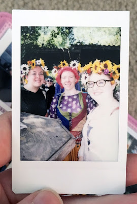 A polaroid of three smiling young white women, rather overexposed, all wearing colorful flower crowns and standing around a high wooden table.
