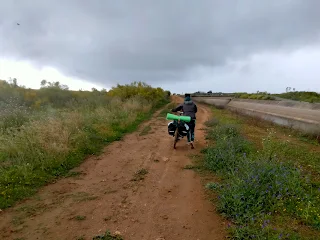 Aim'jie pushes her bike on a wet dirt road, cloudy sky.