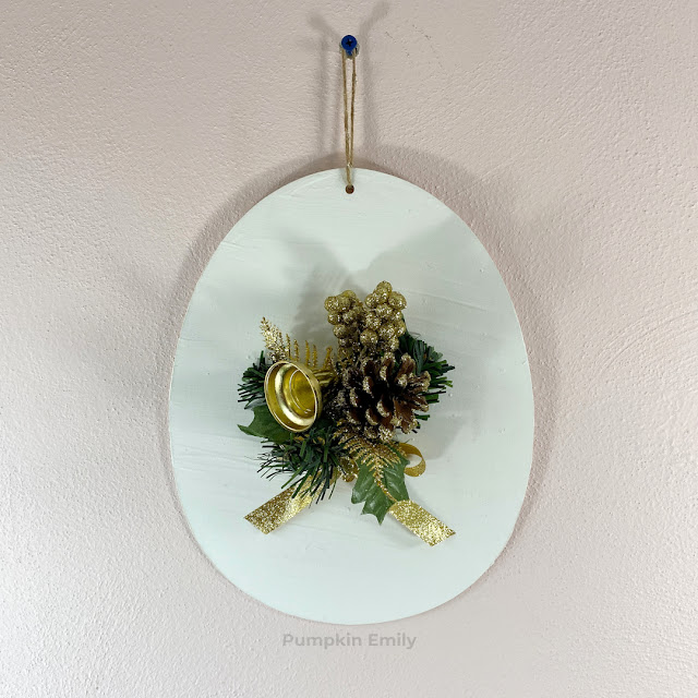 A white wall decoration with a Christmas flower pick and a gold bow.