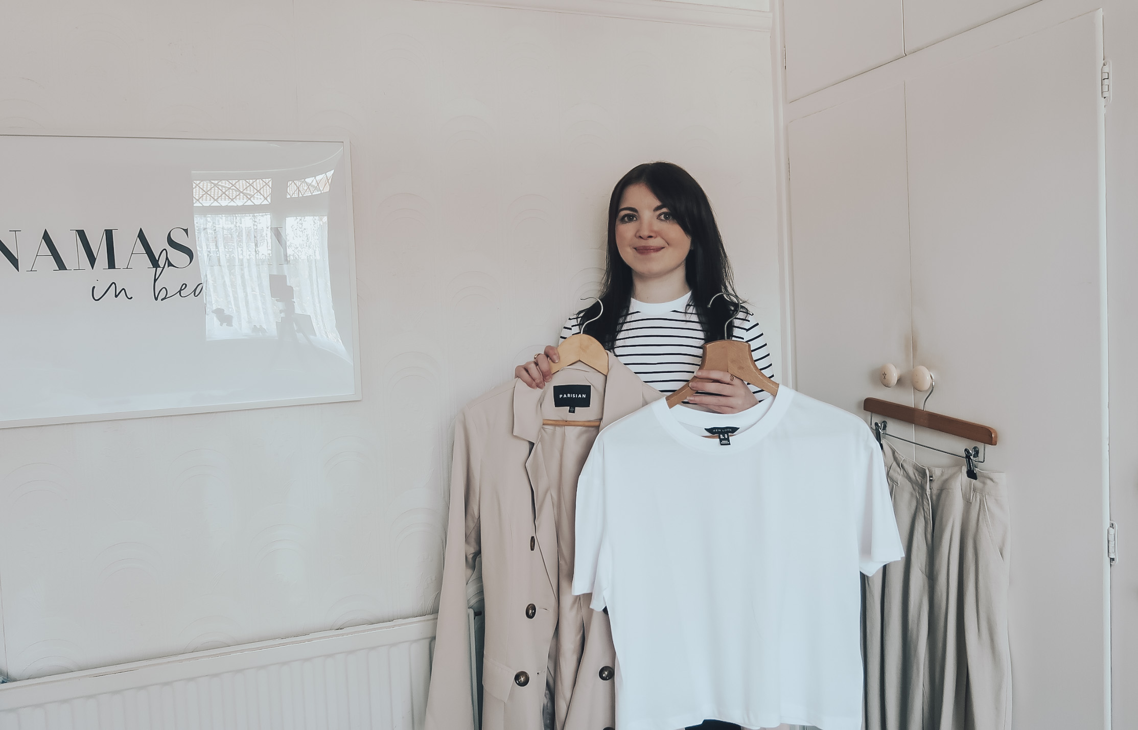 A woman holding a white t-shirt and a beige trench displaying Spring capsule wardrobe.