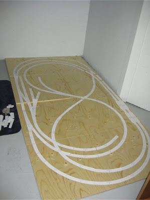 Paper Atlas track template pieces outlining a track design on a 4 x 8 foot sheet of plywood laying on a concrete floor