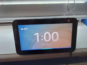 A black Echo Show showing the time 1:00 PM and the weather. The background of the image is showing blinds. The blue Alexa light is showing the at the bottom of the screen.