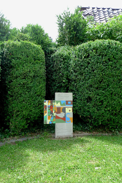 Stèle en béton plantée verticalement dans le sol et peinte avec des fromes colorées aux teintes crayeuses. Un volet en bois est accroché sur la gauche peint avec des formes plus petites