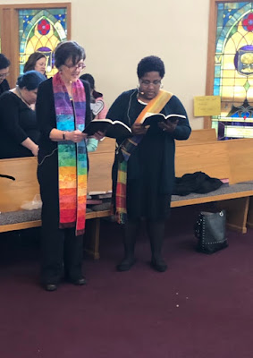 two women, one light-skinned and one dark-skinned, stand side by side, singing