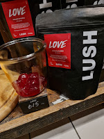 A close up photo of a dark black coffee bag style bag filled with spherical red balls filled with shower gel with a black rectangular label on the bag that says love shower spheres lush in white font on a bright background