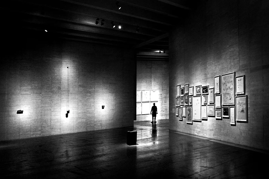 Sala de exposições com o pé direito muito alto. Na parede da esquerda, objectos fortemente iluminados, na da direita quadros pequenos muito aglomerados e em frente, um homem na passagem para outra sala