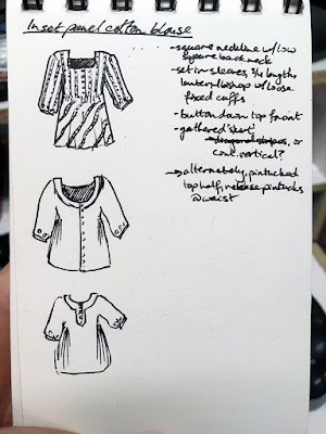 A tiny vertical spiral-bound sketchbook being held in front of a black keyboard. The page is labelled "Inset panel cotton blouse" and has three black pen sketches of button-down shirts, with long, slightly belled sleeves and thin lines describing the pattern direction of the fabric. There are a variety of square and round necks; all are long. A short column of notes in black pen fills half the right side of the page..
