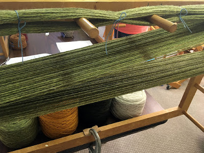 Wide diagonal bands of light green wool, held on a light wooden frame with pegs protruding toward the viewer, and tied with light blue cotton yarn at intervals on the upper pass
