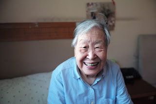 An elderly woman sitting on her bed and smiling.