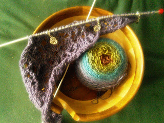 A cake of purple-to-red gradient yarn and an asymmetrical knitted lace triangle, still on the needles.  3 silver rose stitch markers are hooked into the lace.  Both the yarn cake and the lace are inside a wooden yarn bowl.