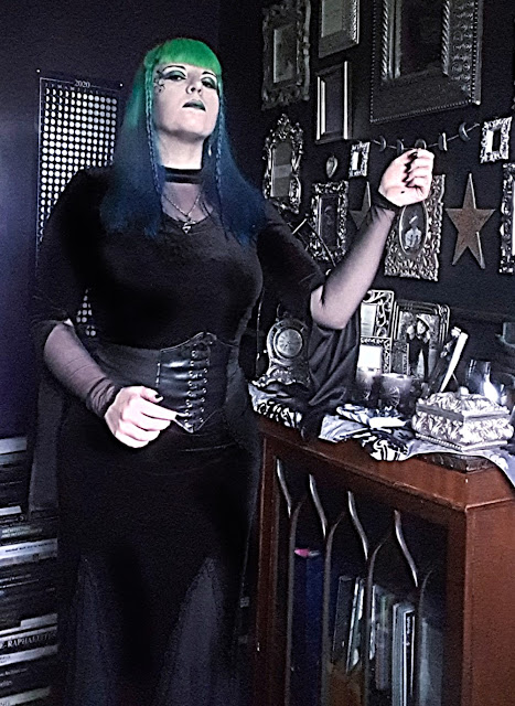 A Gothic woman standing. Behind her is a dark purple wall with a poster of the phases of the moon/moon calendar for 2020. To her left is a black wall with an assortment of ornate silver frames, and a dark-brown wooden cabinet with  glass front of several Gothic arches, on top of which is a silver and black damask table-runner and many ornate silver-coloured items. Behind her to the right is a stack of books on art, architecture and archaeology. She is wearing a close-fitting black velvet top with a v-neck mesh insert at the neck; the sleeves of the blouse have the outer velvet layer split from the wrist to the elbow so the droop down and reveal a mesh inner sleeve. She has an hourglass figure achieved with a corset. She is wearing a faux-lether waist-cincher over her clothes; it laces up at the front, but this is a false closure. She is wearing a black velvet skirt that is close fitting at the top, but flares at the bottom. It has mesh insert panels at the bottom and a 'hankerchief' hem