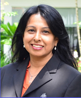 A headshot of Pamela, who is smiling. She looks professional in a charcoal jacket and has a building in the Tropics as her background.