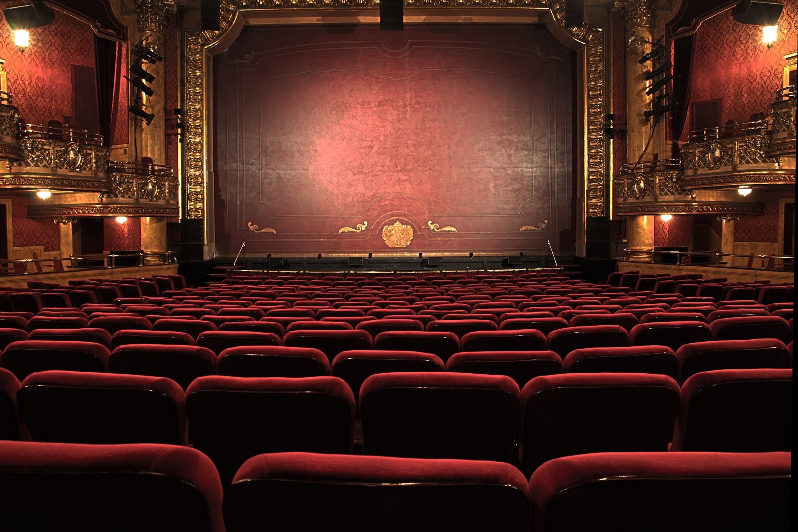 A photo inside a theatre, at the back of the stalls looking over the red seats towards the stage with it's red safetly curtain down