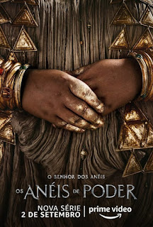 Foto de um par de mãos com manchas de ouro. As mãos são negras, com a direita sobre a esquerda e estão na altura do abdômen de uma pessoa. Ela usa uma roupa com tecido dourado plissado e enfeites triangulares de ouro sobre a roupa, à direita e à esquerda. Nos pulsos veem-se pulseiras de ouro com pedras de esmeraldas e rubis.   Na parte inferior está o título em letras prateadas maiúsculas: "O SENHOR DOS ANÉIS: OS ANÉIS DE PODER". Logo abaixo lê-se em letras brancas maiúsculas: "NOVA SÉRIE 2 DE SETEMBRO". À direita está o logo do Amazon Prime Video.
