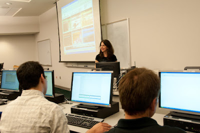 A teacher stands in front of a row of students and their computers.