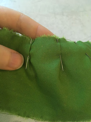 A close-up of three tiny pleats in light lime-green fabric, held with silver pins, and propped up be a white hand with finger and thumb.