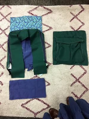 A backpack laid out in several pieces like an engineer's turnaround diagram: back, with blue lining, blue and navy art deco flap, and dark hunter green zipper panels to the left. To the right, the front panel, with a two-sectioned pouch nearly filling the panel. Below the back, a blue rectangle for the base. All laid on a cream rug with a geometric garnet pattern, and with the photographer's long denim skirt and brown boots just showing at lower right.