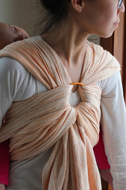 [Image of a tan skin bespectacled Asian woman from nose through torso. She's looking out a window while wearing a finally sleeping toddler on her back in a woven wrap of undyed combed cotton warp and a mercerized orange weft in a modified herringbone pattern. The carry is finished with an orange aluminum Sling Ring in the upper center of the woman's chest. The toddler's sleeping face peeks slightly over the woman's shoulder. Woman has on a cream long-sleeve shirt. They're lit by a cloudy afternoon.]