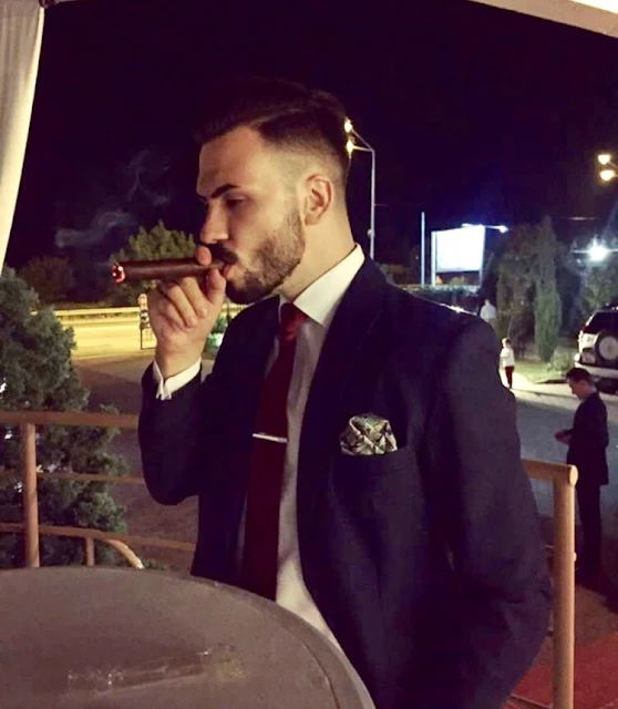 Handsome dark haired man wearing a blue navy blue suit from the side view smoking a cigar outside sitting
