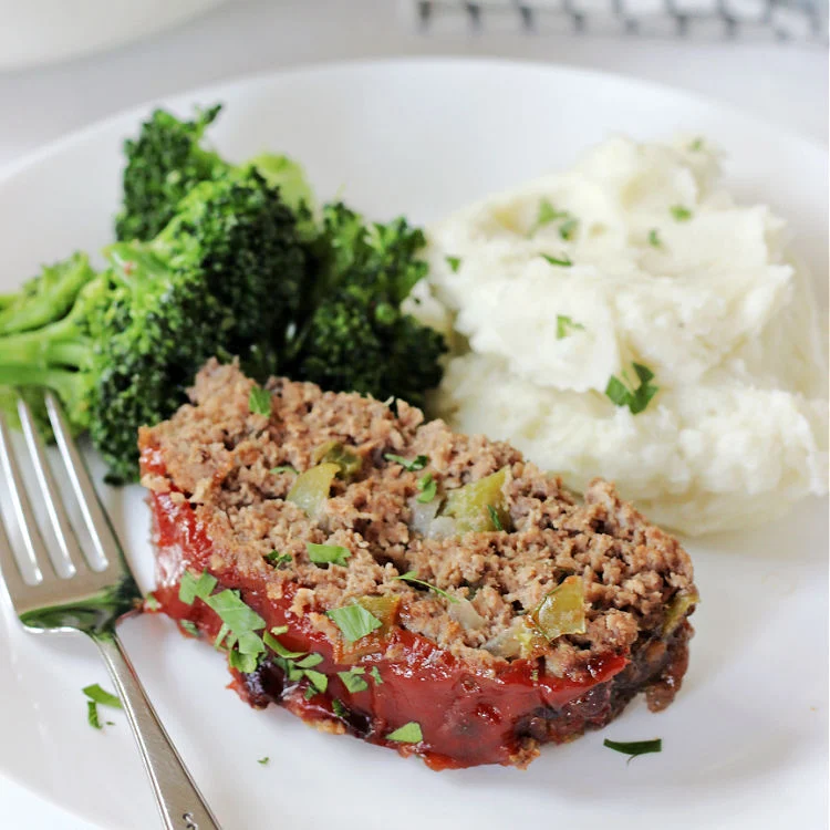 A slice of easy homestyle meatloaf recipe on a plate with steamed broccoli and mashed potatoes and a fork ready to eat