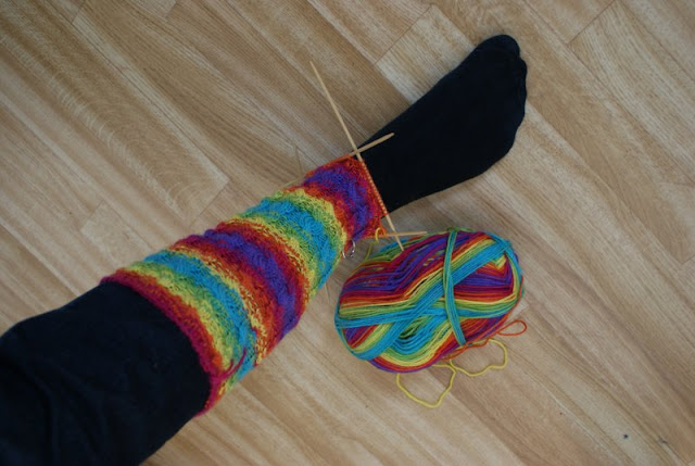 Regnbågsfärgat strumpskaft stickad med flätor. Strumpan visas på ett ben. / Rainbow coloured cable-knitted cuff. The sock is showed on a leg