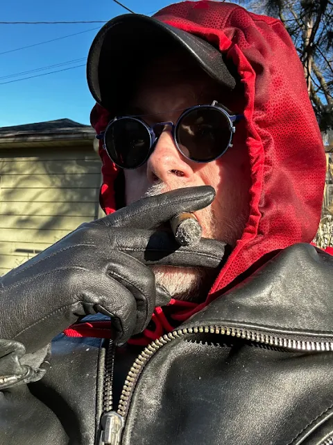 3/12 man standing outside wearing sunglasses with a red hoodie pulled over head and they black leather jacket over top of that gloves holding cigar in your mouth