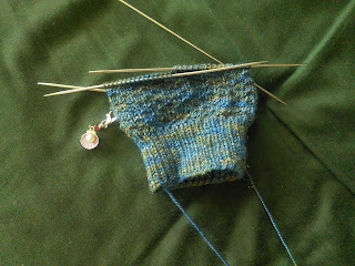 The leg of a textured sock on double-pointed needles.  The yarn is a blue and gold variegated yarn with a small amount of sparkle. There is a a stitch marker clipped into the work, the marker looks like a shell with a purl inside. 