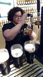 Woman in a black t-shirt pouring a Guiness beer from the tap. Behind her is a wall of beer bottles. In front of her is a row of 3 partially poured pints of Guiness beer.
