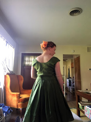 A young redheaded woman with her hair braided up and flowers stuck in it, standing with her back to the camera in a living room cluttered with furniture and sewing materials. She's wearing a green dress with a loose, sleeveless top and a pleated knee-length skirt.