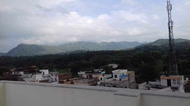 "A panoramic view of a mountain range with a cloudy sky. Below, there are houses and a tall cell tower."