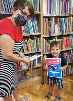 Zdjęcie chłopiec w granatowej koszulce spodenkach w niebieskie paski odbiera dyplom, dodatki, zakładki z rąk bibliotekarki w tle regały z książkami