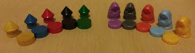 On the left are the pieces from Clans: wooden pieces resembling round huts with conical roofs, in each of the five colours. The pieces use basic, bold primary colours. On the right are pieces from Fae, plastic figurines which resemble small hooded figures with their hands clasped in front of them. The colours are more muted. In front of each piece on both sides are the scoring discs for the corresponding colour. The ones from Clans are wooden, and are slightly larger than the ones from Fae, which are plastic.