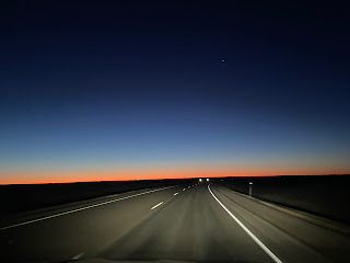 a highway lit up by headlights and a straight lined horizon glowing orange that fades to dark blue sky